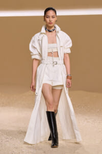 Model on a runway wearing an all-white utility set: long open coat, cropped top, high-waisted shorts, and tall black boots.