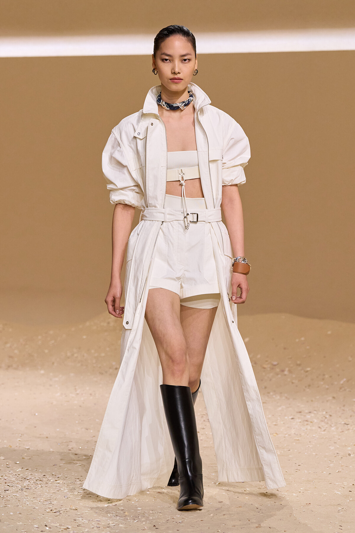 Model on a runway wearing an all-white utility set: long open coat, cropped top, high-waisted shorts, and tall black boots.