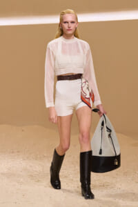 Model walking the runway in a pale pink sheer cropped top over a white bralette, white high-waisted shorts, and knee-high black boots, carrying a large two-tone bag and scarf.
