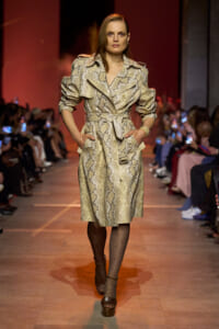 Female model walks a runway wearing a beige snakeskin-pattern trench coat with a belt and platform sandals, hands in pockets.