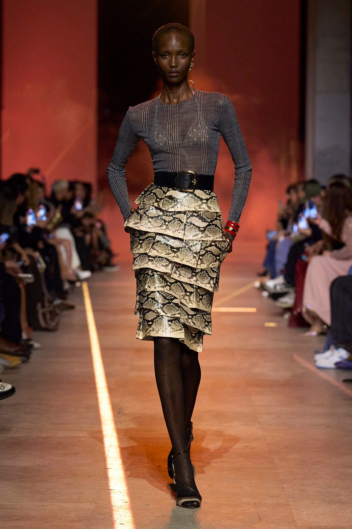 Model walking the runway in a gray, fitted long-sleeve top with a multi-layered snakeskin-print peplum skirt and wide black belt.