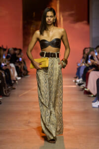 Model walking runway in a black strapless top, snake-pattern wide-leg pants, and a yellow clutch.