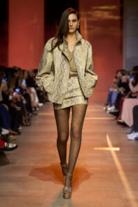 Model walks the runway in a snakeskin-print beige blazer and matching mini skirt, with an olive top, sheer polka-dot tights, and beige platform sandals; audience in the background.