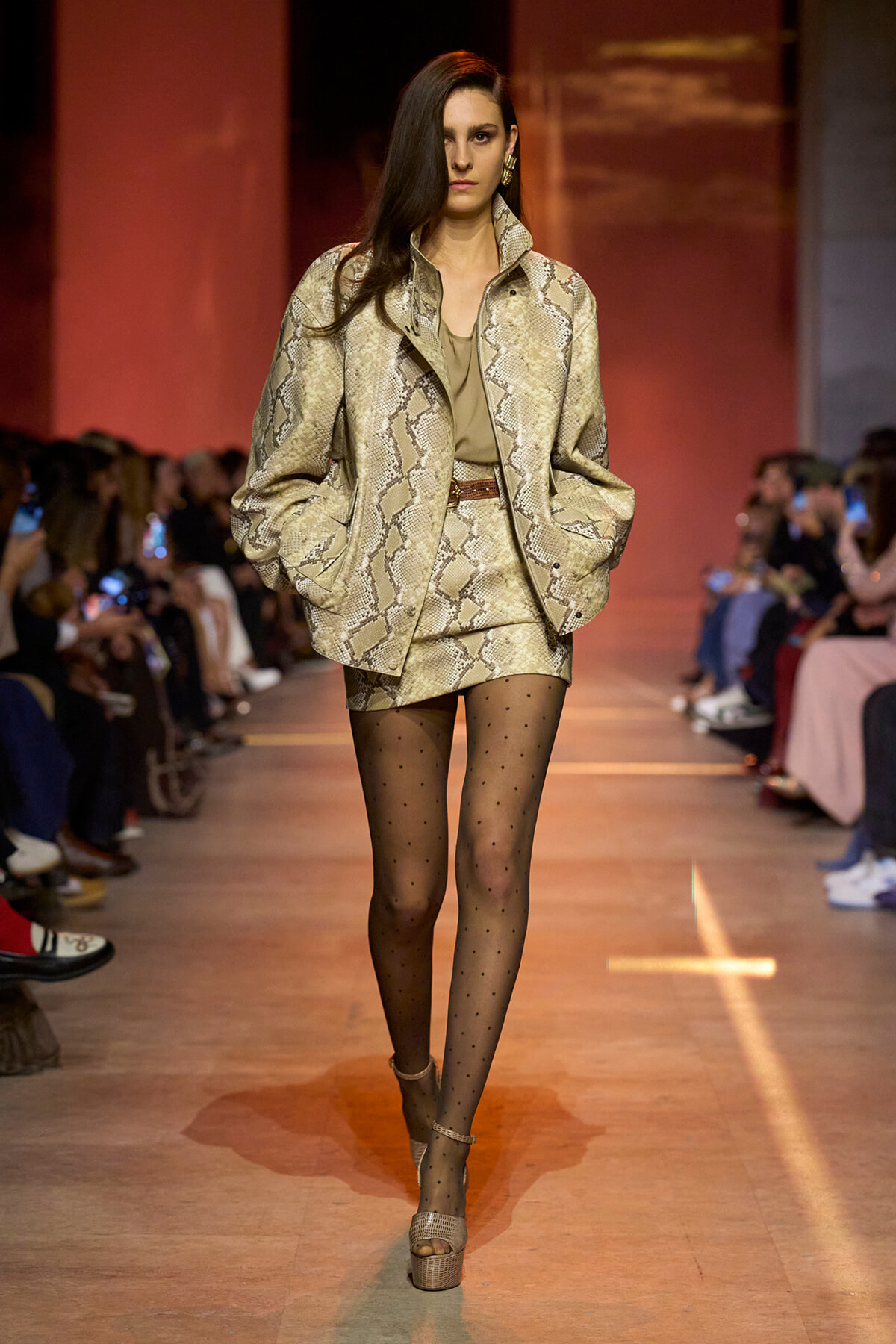 Model walks the runway in a snakeskin-print beige blazer and matching mini skirt, with an olive top, sheer polka-dot tights, and beige platform sandals; audience in the background.