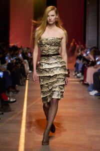 Model walking runway in a strapless snakeskin-print layered dress with a black belt and gold earrings; audience on sides.