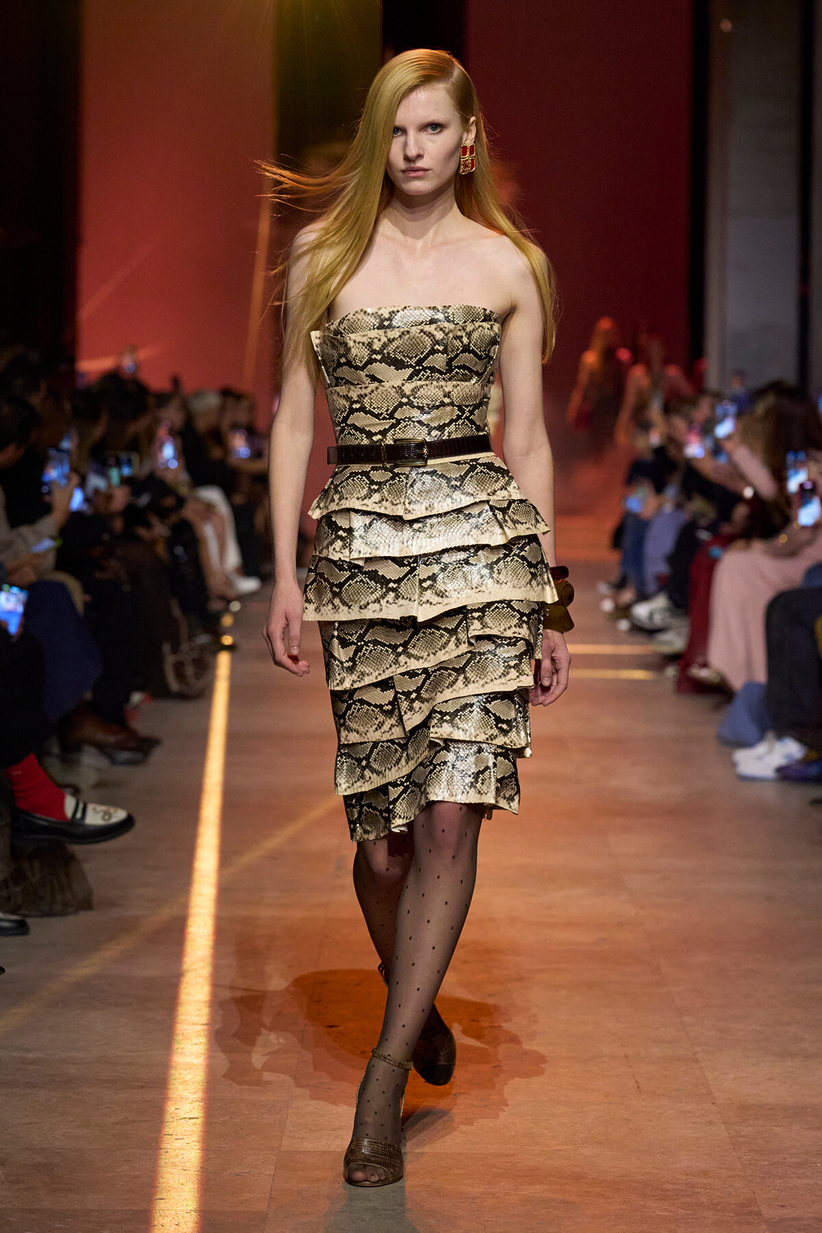Model walking runway in a strapless snakeskin-print layered dress with a black belt and gold earrings; audience on sides.