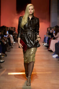 Model walks the runway in a black leather belted jacket with a gold sequin midi skirt and red clutch.