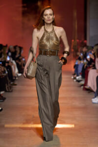Model walks a fashion runway in a bronze metallic halter top, gray wide-leg trousers, a wide brown belt, chunky bracelets, and a textured beige handbag.