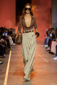 Model walking runway in a brown plunging top with wide beige pants and a brown belt, carrying a textured handbag and wearing oversized sunglasses.