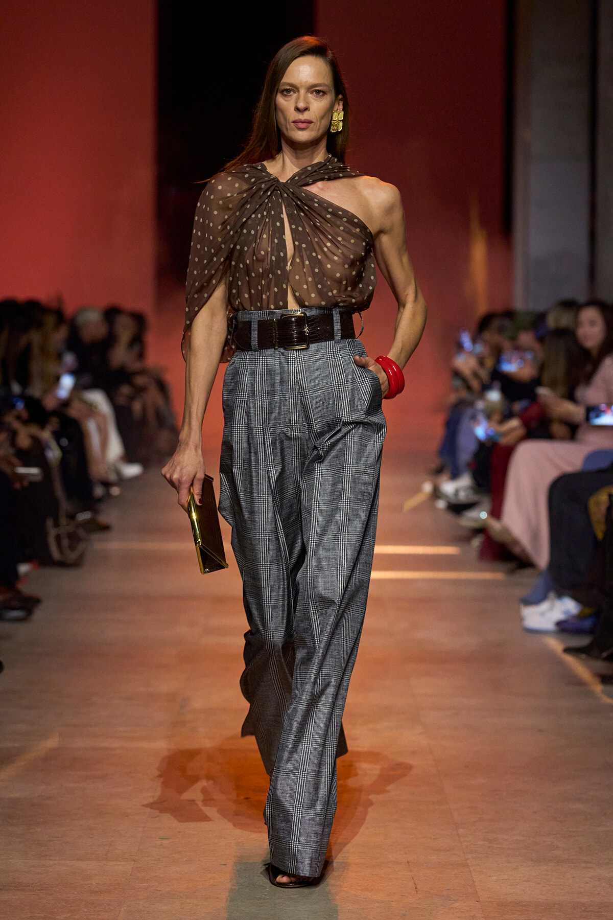 Model on a runway wearing a sheer brown polka-dot top, gray plaid wide-leg pants, and a wide black belt, holding a gold clutch.