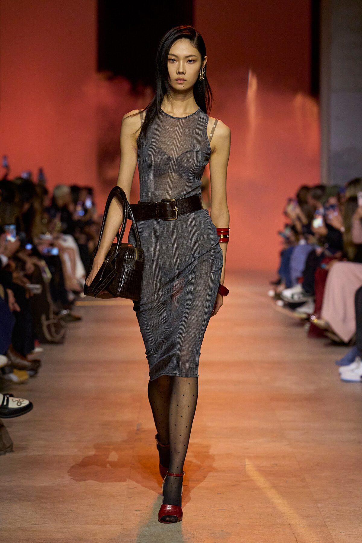 Model walking a runway in a plaid gray sleeveless dress with a wide dark belt and black handbag, red accessories, and dotted tights.