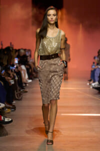 Model walking the runway in a gold sequin sleeveless top tucked into a beige checkered knee-length skirt with a wide brown belt and brown heels, during a fashion show.