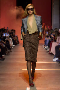 Model on a runway wearing a gray plaid blazer over a tan draped top and a brown polka-dot skirt, with red shoes and large sunglasses.