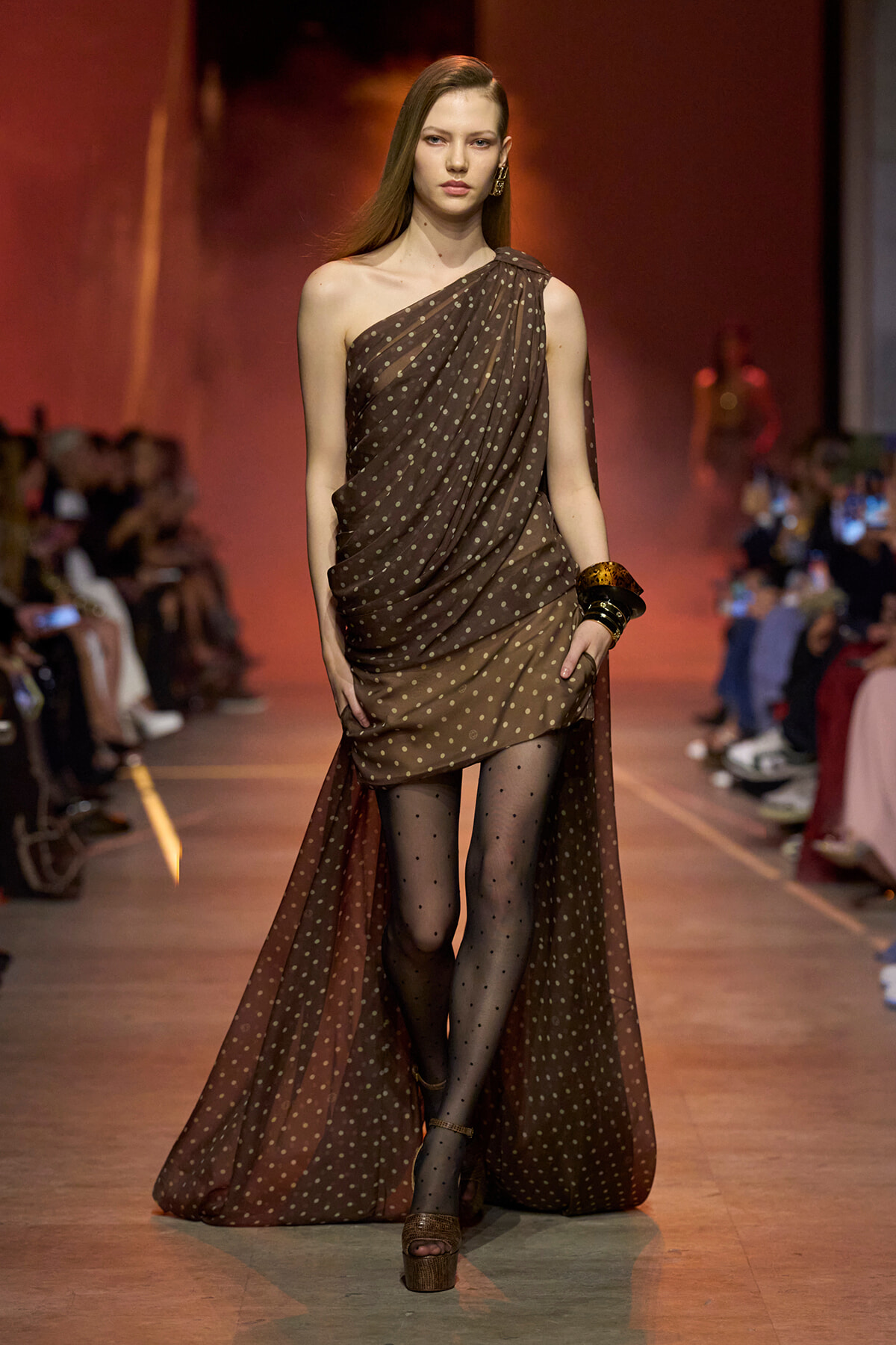 Female model walks the runway in a brown, polka-dot, one-shoulder sheer gown with a long draped cape and polka-dot tights, paired with platform sandals and stacked bangles.