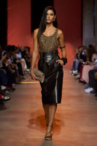 Model walking a runway in a metallic beaded bronze top and high-waisted black leather midi skirt, holding a beige clutch.