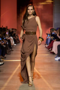 Female model walks a brown one-shoulder draped gown with a wide brown belt down a runway, holding a small clutch.