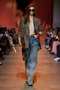 Model on a runway wearing a gray double-breasted blazer, beige blouse, and oversized sunglasses with wide-leg denim jeans.
