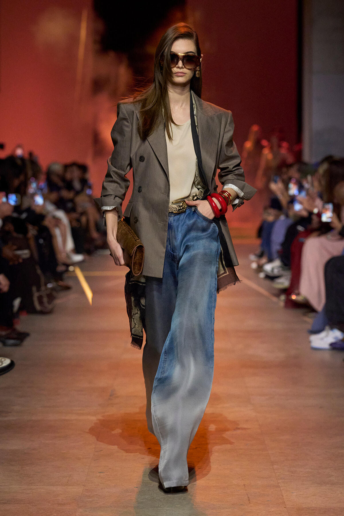Model on a runway wearing a gray double-breasted blazer, beige blouse, and oversized sunglasses with wide-leg denim jeans.