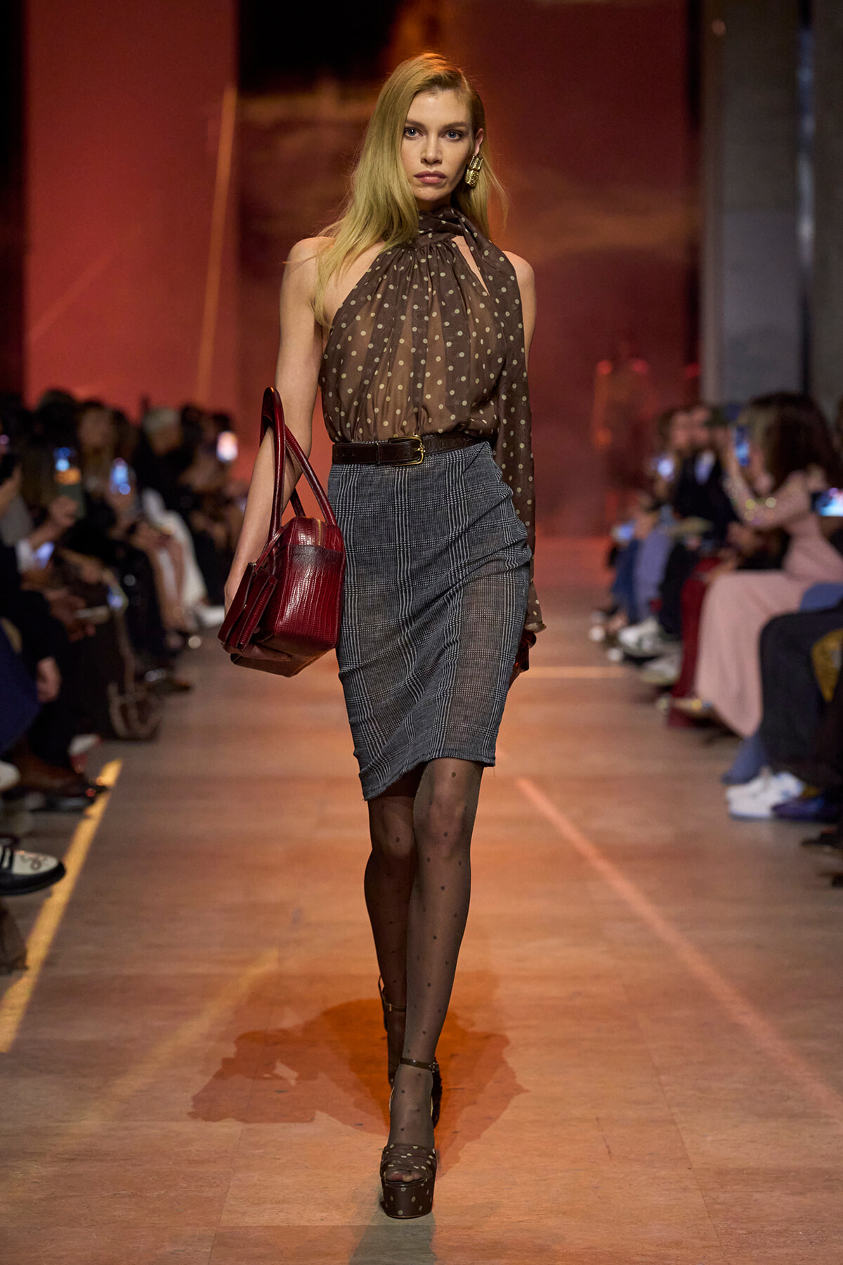 Model walking runway in a brown polka-dot halter blouse, gray plaid pencil skirt, and red handbag, with sheer polka-dot tights and sandals.