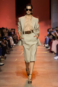 Model walking a runway in a beige tailored suit with a draped pale top, belt, and heeled sandals; large dark sunglasses and gold accessories; warm orange backdrop.