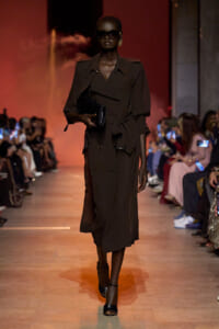 Model walks the runway in a dark belted trench coat, oversized shoulders, black clutch and sunglasses.