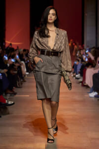 Model walking a fashion runway in a taupe checkered blouse with deep V-neck and a high-waisted gray knee-length skirt, cinched with a wide belt, holding a snakeskin clutch, audience along the sides.