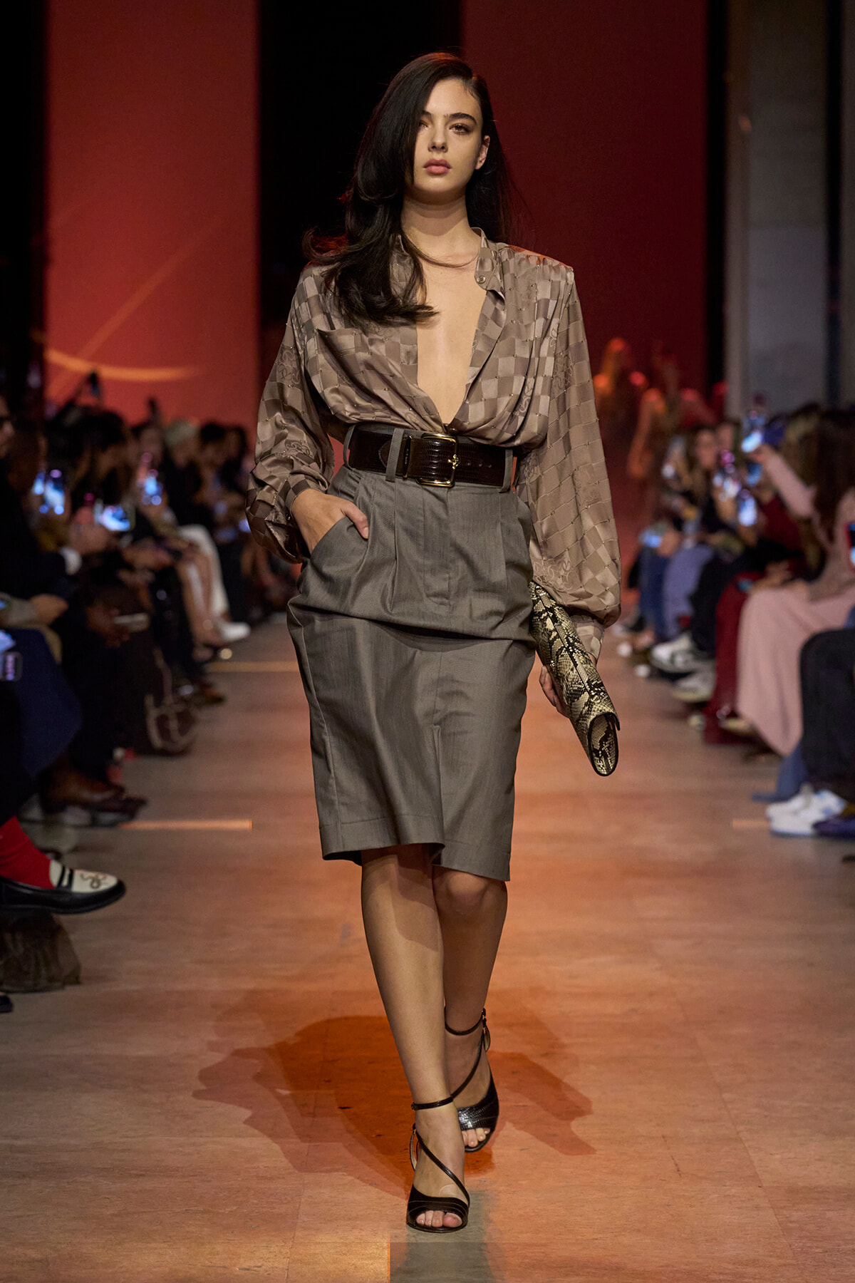 Model walking a fashion runway in a taupe checkered blouse with deep V-neck and a high-waisted gray knee-length skirt, cinched with a wide belt, holding a snakeskin clutch, audience along the sides.