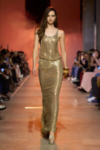Model walks the runway in a gold sequined tank top and matching long skirt, accessorized with stacked bangles.