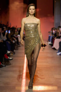 Model walks the runway in a gold sequined strapless dress with an asymmetric drape, paired with dotted sheer tights and bracelets.