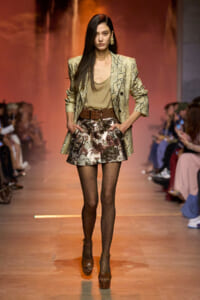 Model walks a runway in a snake-print blazer, taupe top, metallic floral shorts, and brown platform heels with sheer dotted tights.