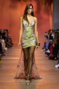 Runway model walks in a strapless, abstract floral mini dress with a sheer floral cape and dotted tights; wearing chunky bracelets and silver sandals.