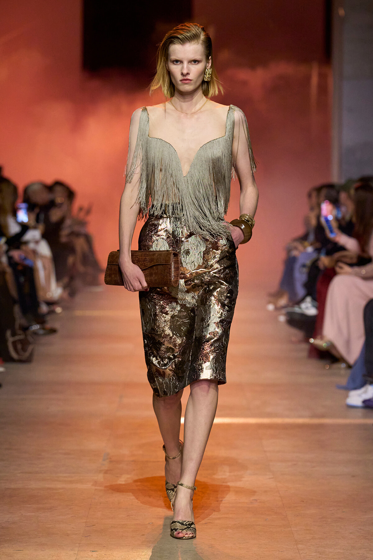 Model on runway wearing a pale green fringe top with a metallic brocade skirt, holding a brown clutch.