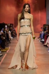 Model walks the runway in a strapless nude gown with snake-print bands and a high slit, audience blurred in the background.