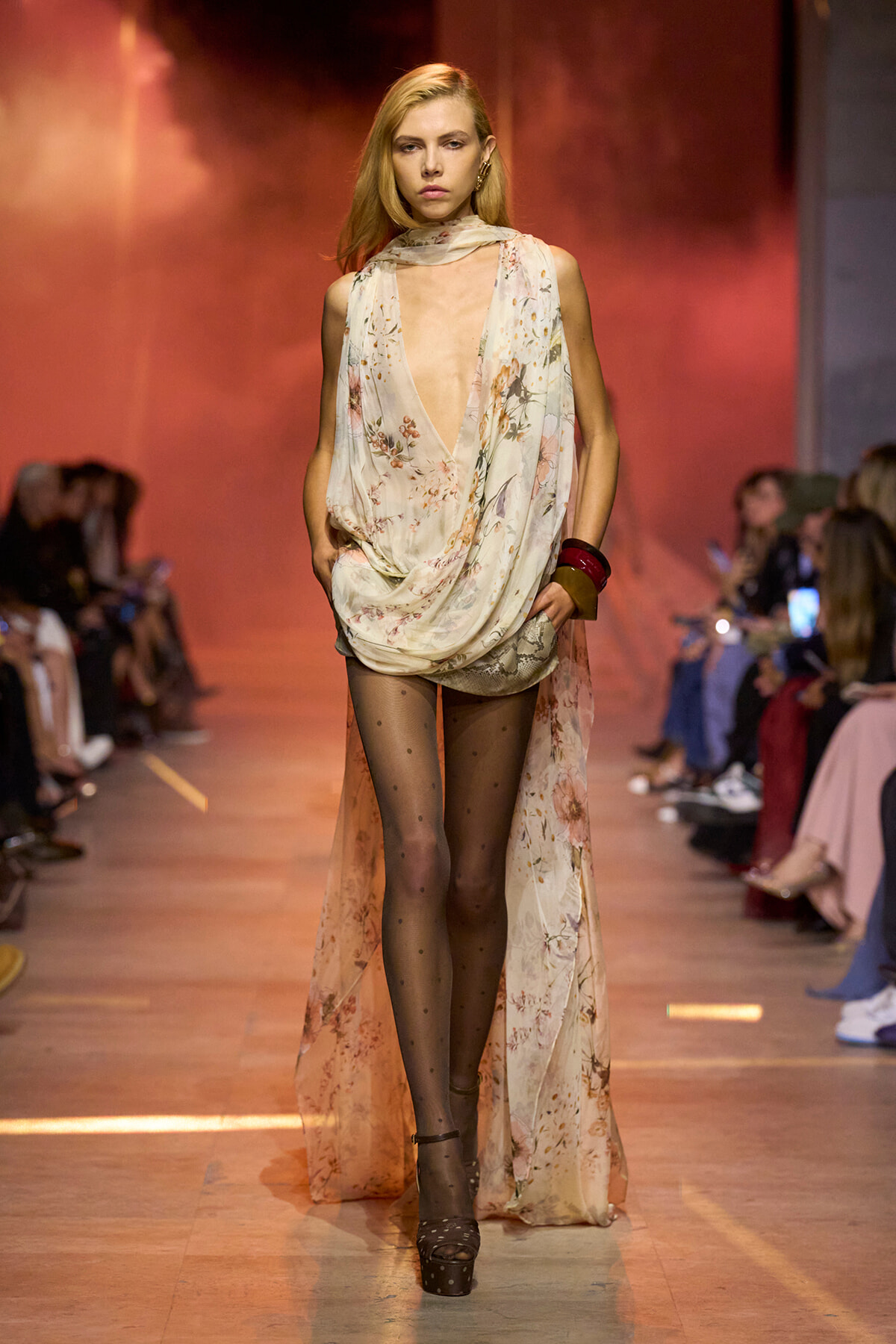 Model walks a floral sheer draped gown with a deep V neckline on a runway, paired with dotted sheer tights and platform sandals, wearing bold bracelets.