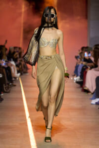 Model on a fashion runway wearing a snakeskin bandeau top and a draped taupe skirt with a high slit, plus a sheer scarf and oversized sunglasses.