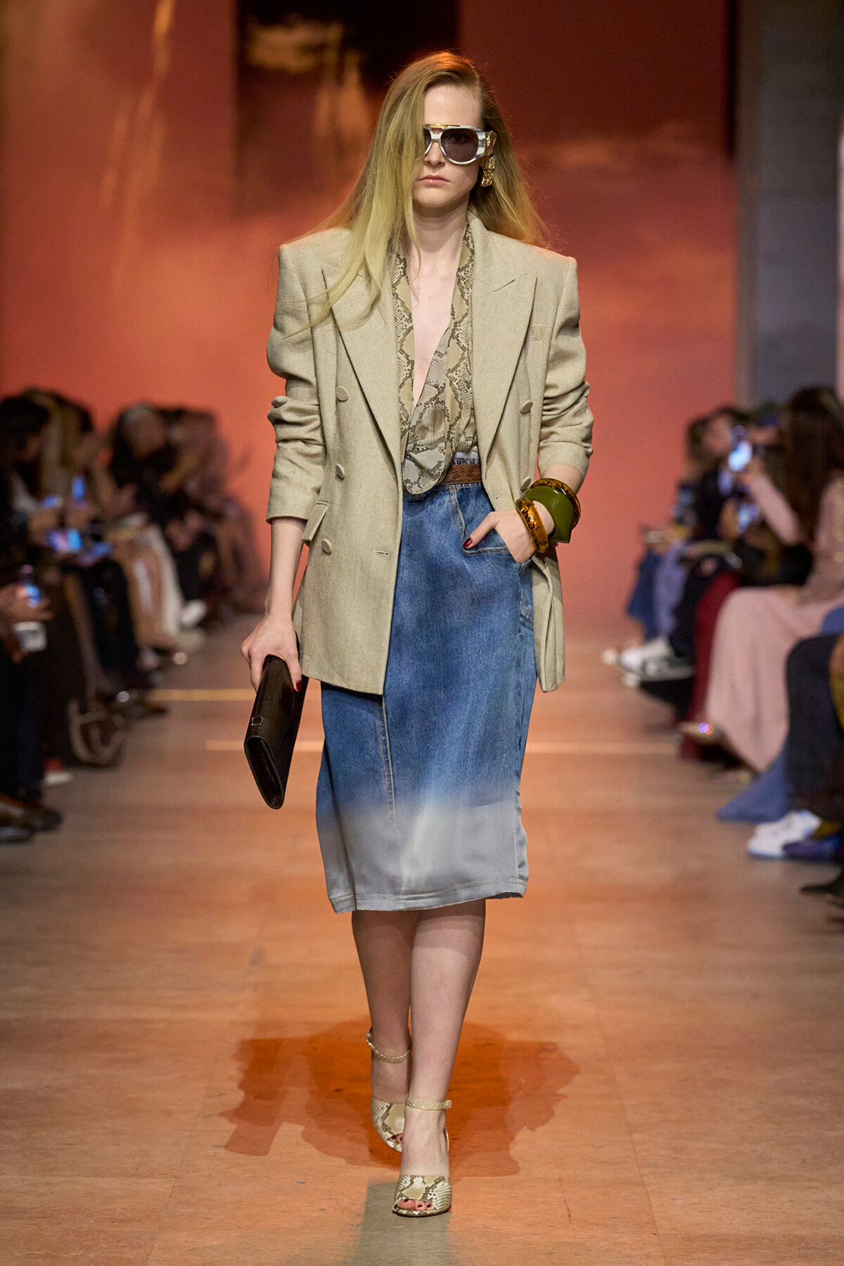 Runway model wearing a beige oversized blazer, snake-print blouse, and a gradient denim skirt, carrying a clutch and wearing large sunglasses.