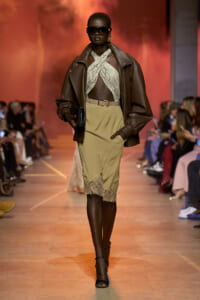 Model walks the runway in a brown leather jacket over a snakeskin halter top and a beige lace-trimmed skirt, with oversized sunglasses and black heels.