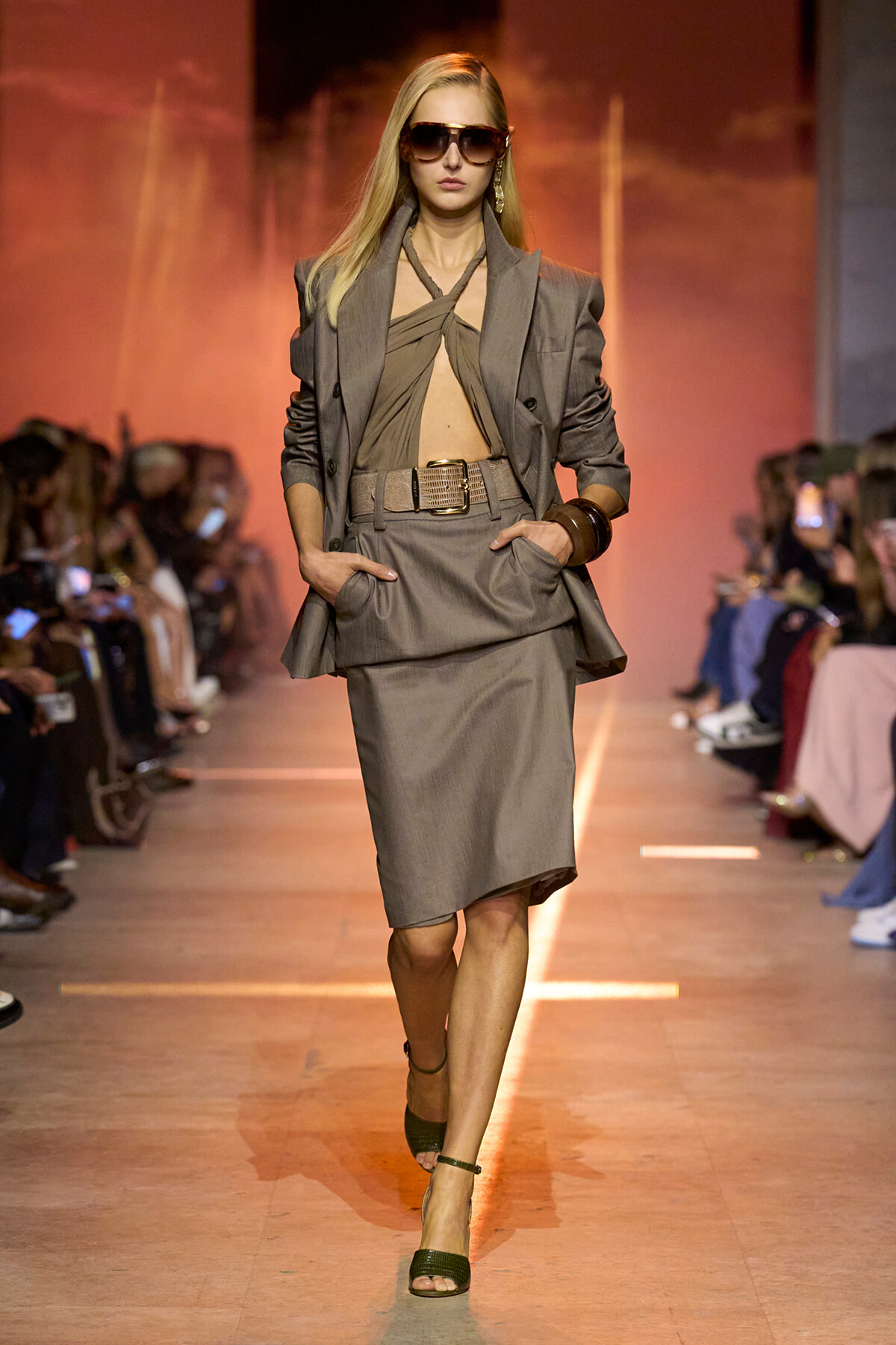 Model walks the runway in a taupe-gray business suit: structured blazer over a knotted top, matching pencil skirt, belt, sunglasses, and open-toe heels.
