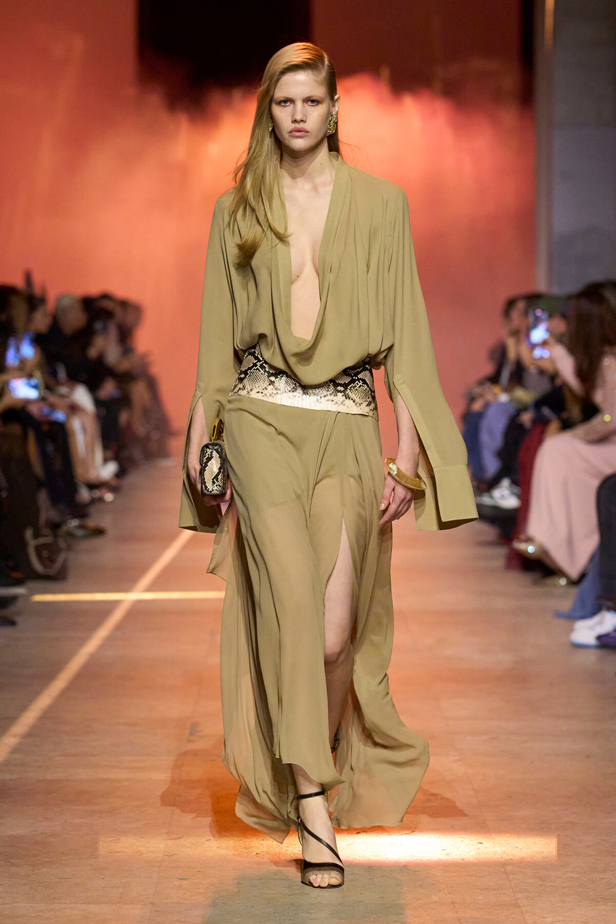 Model on a runway wearing an olive draped jumpsuit with a deep plunging neckline, snakeskin belt, and black strappy heels, orange backdrop behind.