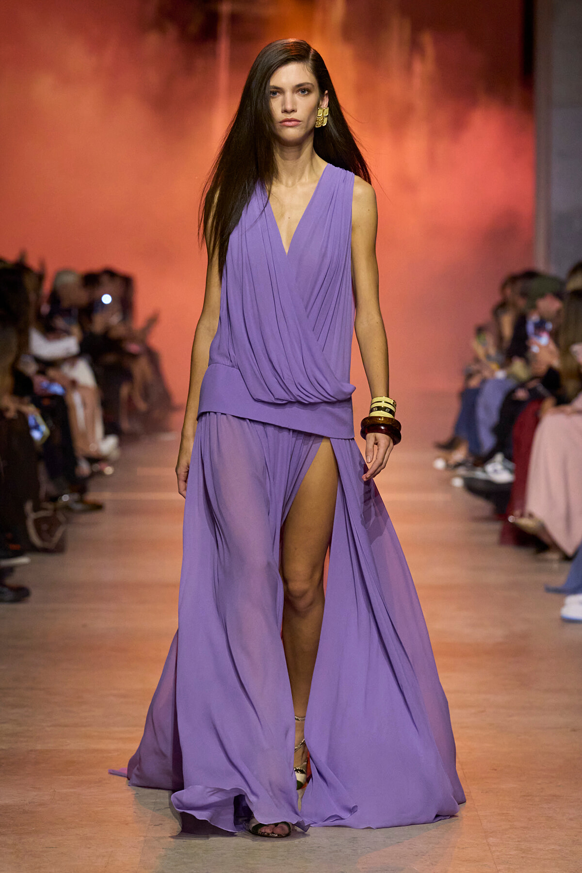 Model walks the runway in a lavender draped sleeveless gown with a high slit, accessorized with gold earrings and bangles.