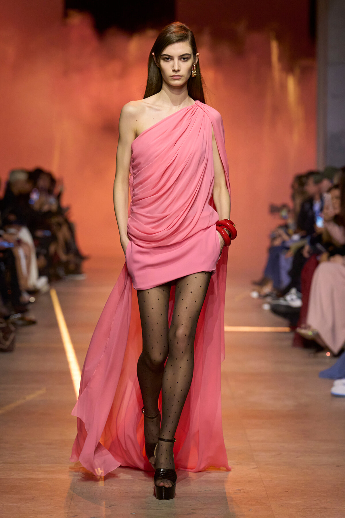 Model walking the runway in a pink draped one-shoulder dress with a long sheer cape, black dotted tights, and platform sandals.