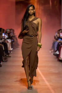 Model walks a brown draped one-shoulder dress on the runway, holding a metallic clutch and wearing bamboo bangles and black sandals on a warm-lit catwalk.