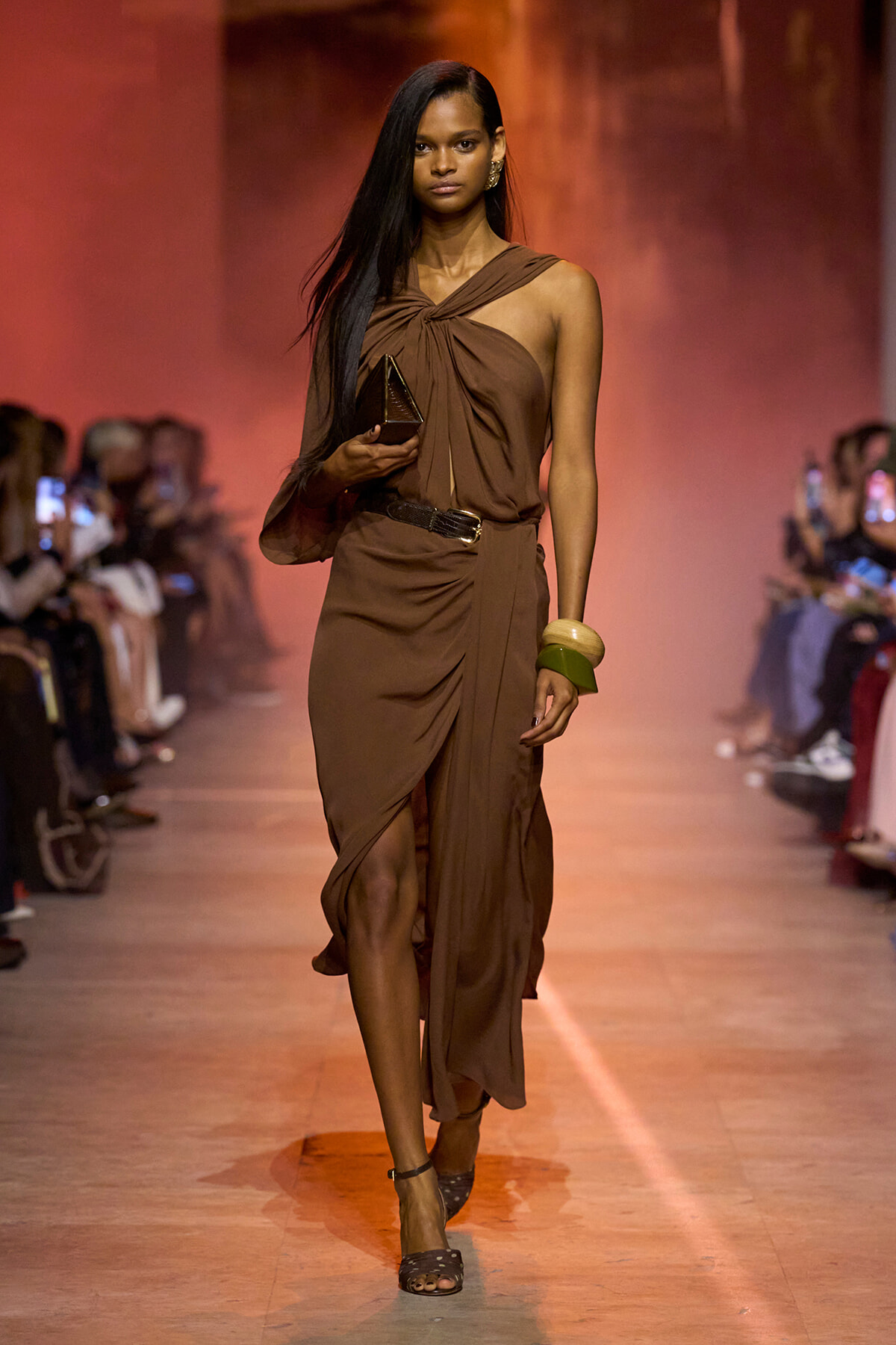 Model walks a brown draped one-shoulder dress on the runway, holding a metallic clutch and wearing bamboo bangles and black sandals on a warm-lit catwalk.