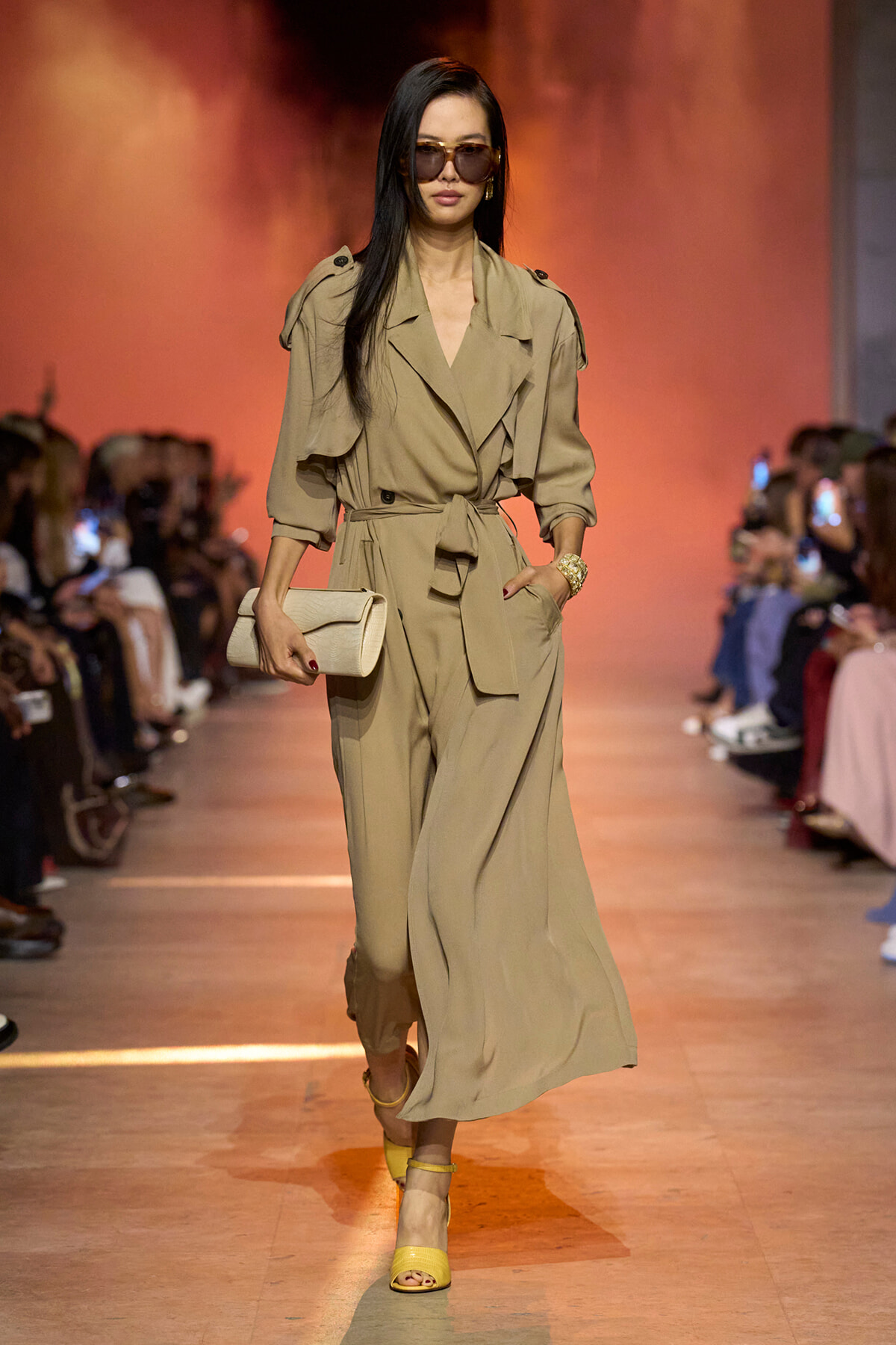 Model walks the runway in a tan belted trench-style jumpsuit, oversized sunglasses, and a beige clutch.