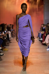 Runway model wearing a lavender one-shoulder, draped gown with a waist cutout and ruched skirt, accessorized with brown bangles.