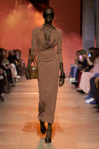 Model walks the runway in a taupe draped maxi dress, oversized sunglasses, and stacked bracelets, carrying a brown handbag.