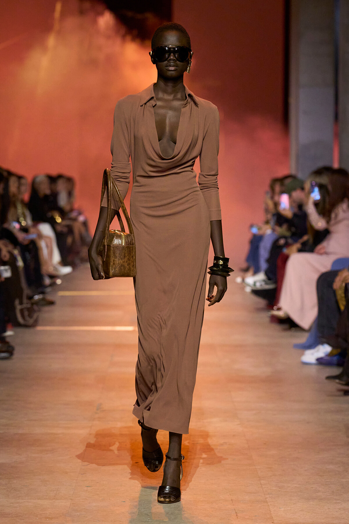 Model walks the runway in a taupe draped maxi dress, oversized sunglasses, and stacked bracelets, carrying a brown handbag.