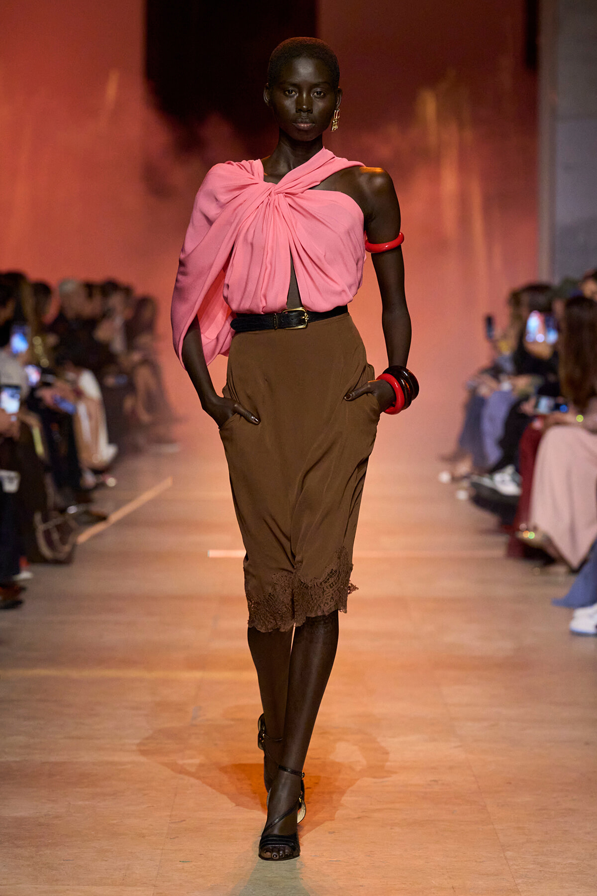 Model walking a runway in a pink draped one-shoulder top and brown midi skirt with lace hem, accessorized with red bangles and black heels.