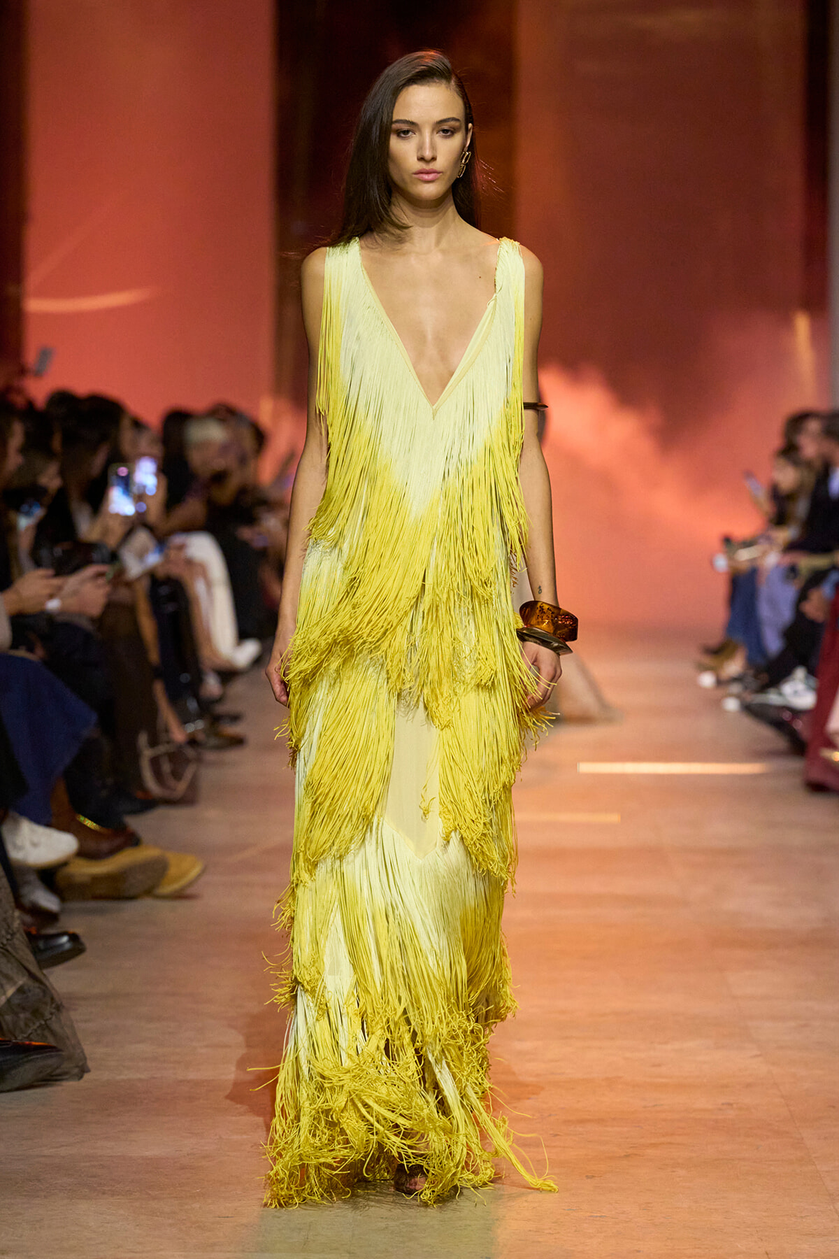 Model walks a runway in a long yellow fringe gown with a deep V neckline.