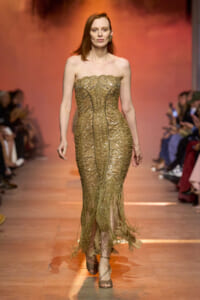 Model walking the runway in a strapless, gold beaded gown with intricate pattern and fringe at the hem, during a fashion show.
